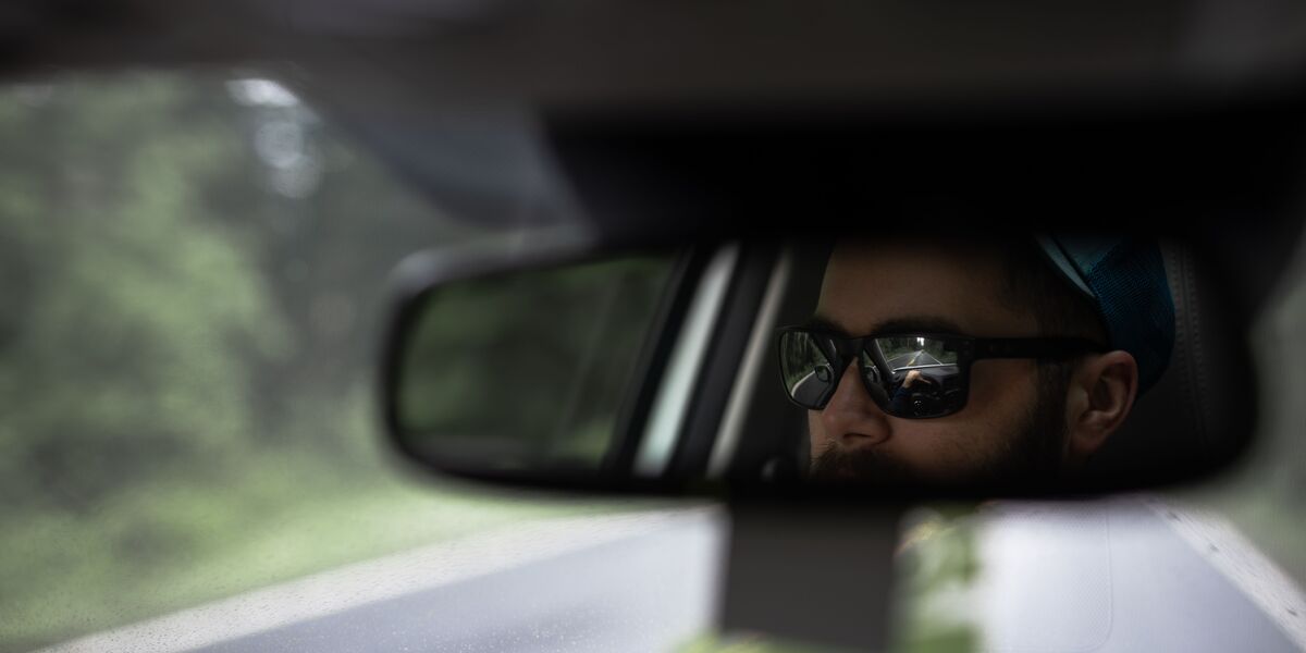 Driver wearing sunglasses seen in the rear-view mirror on a country road