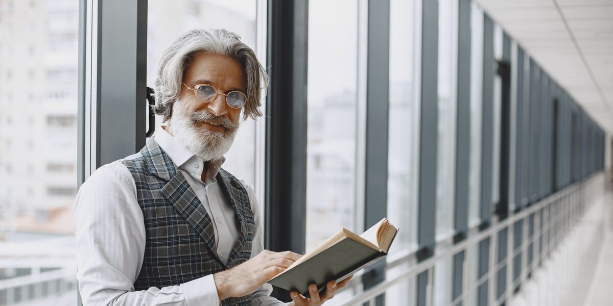 Older man with grey hair and a beard holding an open book and looking up through his glasses